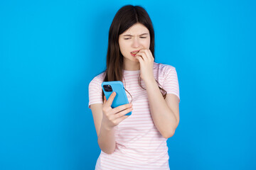 Portrait of pretty frightened young beautiful Caucasian woman wearing stripped T-shirt over blue wall chatting biting nails after reading some scary news on her smartphone.