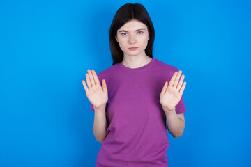 Serious young beautiful Caucasian woman wearing purple T-shirt over blue wall pulls palms towards camera, makes stop gesture, asks to control your emotions and not be nervous