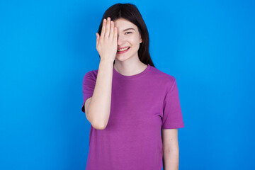 young beautiful Caucasian woman wearing purple T-shirt over blue wall covering one eye with her hand, confident smile on face and surprise emotion.