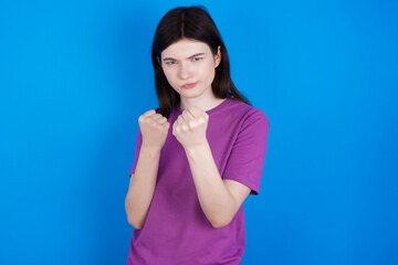 young beautiful Caucasian woman wearing purple T-shirt over blue wall Ready to fight with fist defense gesture, angry and upset face, afraid of problem.