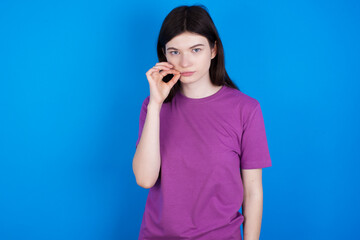 young beautiful Caucasian woman wearing purple T-shirt over blue wall mouth and lips shut as zip with fingers. Secret and silent, taboo talking.