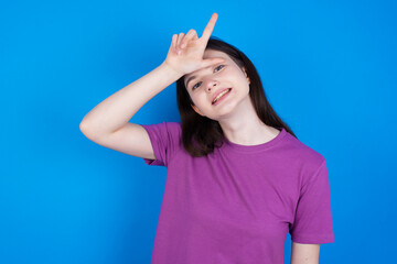 young beautiful Caucasian woman wearing purple T-shirt over blue wall making fun of people with fingers on forehead doing loser gesture mocking and insulting.