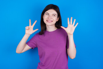Obraz na płótnie Canvas young beautiful Caucasian woman wearing purple T-shirt over blue wall showing and pointing up with fingers number eight while smiling confident and happy.