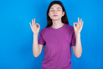 young beautiful Caucasian woman wearing purple T-shirt over blue wall relax and smiling with eyes closed doing meditation gesture with fingers. Yoga concept.