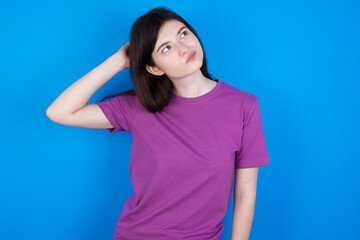 young beautiful Caucasian woman wearing purple T-shirt over blue wall confuse and wonder about question. Uncertain with doubt, thinking with hand on head. Pensive concept.