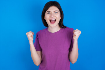 young beautiful Caucasian woman wearing purple T-shirt over blue wall celebrating surprised and amazed for success with arms raised and open eyes. Winner concept.