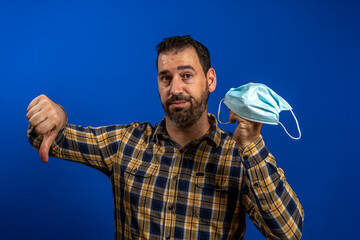 Bearded man in a yellow plaid shirt holding a surgical mask in one hand and making the no OK gesture with the other hand on blue studio background