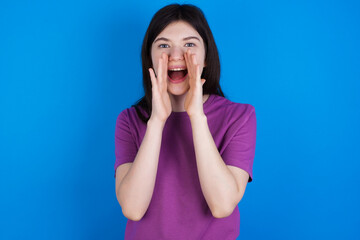 young beautiful Caucasian woman wearing purple T-shirt over blue wall shouting excited to front.