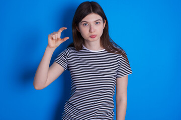 young beautiful Caucasian woman wearing pink jacket over blue wall purses lip and gestures with hand, shows something very little.