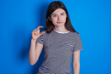 Fototapeta premium Displeased young beautiful Caucasian woman wearing stripped T-shirt over blue wall shapes little hand sign demonstrates something not very big. Body language concept.