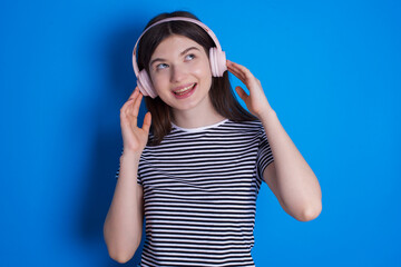 Joyful young beautiful Caucasian woman wearing stripped T-shirt over blue wall sings song keeps hand near mouth as if microphone listens favorite playlist via headphones
