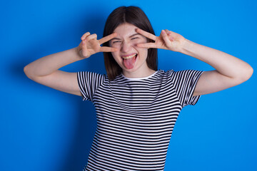 Cheerful positive young beautiful Caucasian woman wearing stripped T-shirt over blue wall shows v-sign near eyes open mouth