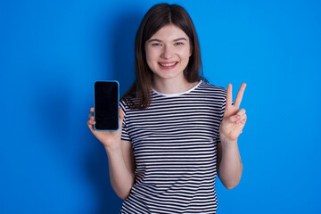 young beautiful Caucasian woman wearing stripped T-shirt over blue wall holding modern device showing v-sign