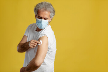 Old Caucasian patient showing vaccinated arm after vaccine injection, yellow background.