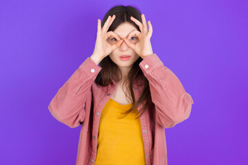 Playful excited young beautiful Caucasian woman wearing casual clothes over purple wall showing Ok sign with both hands on eyes, pretending to wear spectacles.