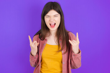Born to rock this world. Joyful young beautiful Caucasian woman wearing casual clothes over purple wall screaming out loud and showing with raised arms horns or rock gesture.