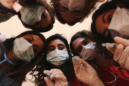 Group Of Young Women Healthcare Workers / Doctors / Medical Students / Medicine Interns With Face Masks, Syringes, Stethoscopes, Surgical Gloves Looking Down On Patient