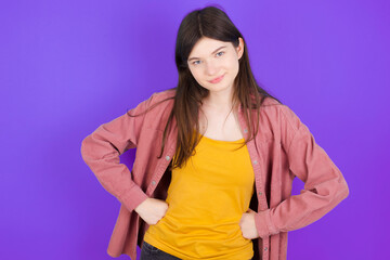 Funny frustrated young beautiful Caucasian woman wearing casual clothes over purple wall holding hands on waist and silly looking at awkward situation.