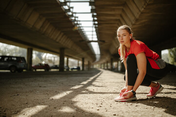 Side view of fit young woman tying shoe laces while standing under the bridge, ready for running.
