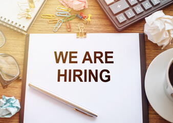 On a wooden table there is an office sheet of paper with the text WE'RE HIRING . Business workspace with calculator, glasses, pen, crumpled paper and cup of coffee.