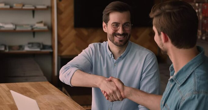 Millennial male colleagues finish collaborative project in office shake hands express gratitude for successful cooperation. Client and manager use laptop make deal accomplish negotiations handshaking