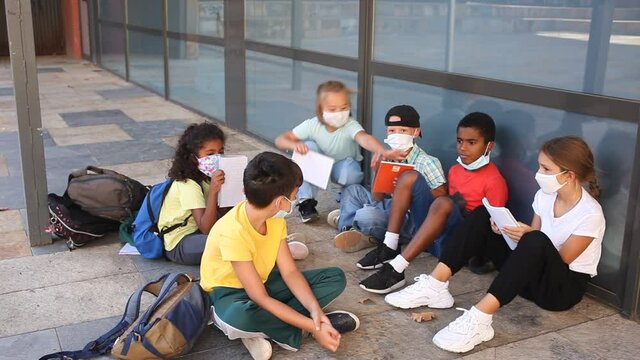 Group of preteen children in face masks resting outdoors in schoolyard during break in lessons. Back school concept during pandemic. High quality FullHD footage