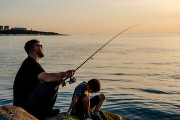 A fisherman sits on a rock by the sea with a fishing rod at sunset. Single sea fishing. Day at the beach. Copy space for text