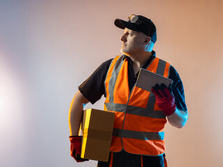 Warehouse worker with a cardboard box. Man works in warehouse or logistics sorting center. He is involved in fulfillment process. Warehouse worker in orange vest. Fulfillment center employee career. - obrazy, fototapety, plakaty