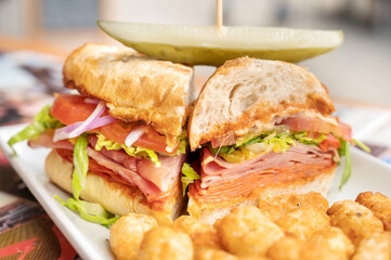 A spicy Italian sandwich with ham, salami, peppers, onions, tomatoes, pickle, cheese, and lettuce.  Served at an out door restaurant cafe.