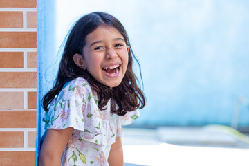 Uma criança, menina, brasileira dos cabelos pretos e lisos, alegre, sorrindo, encostada no muro.
