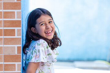 Uma criança, menina, brasileira dos cabelos pretos e lisos, alegre, sorrindo, encostada no muro.