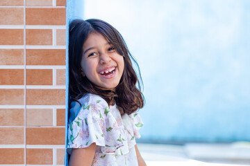 Uma criança, menina, brasileira dos cabelos pretos e lisos, alegre, sorrindo, encostada no muro.