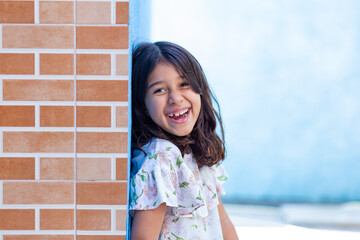 Uma criança, menina, brasileira dos cabelos pretos e lisos, alegre, sorrindo, encostada no muro.