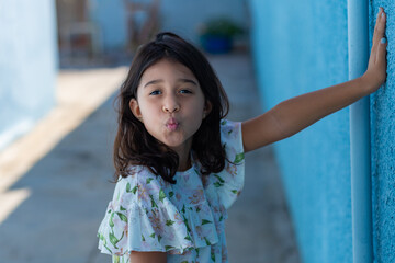 Uma criança, menina, brasileira dos cabelos pretos e lisos, fazendo biquinho encostada no muro pintado de azul.