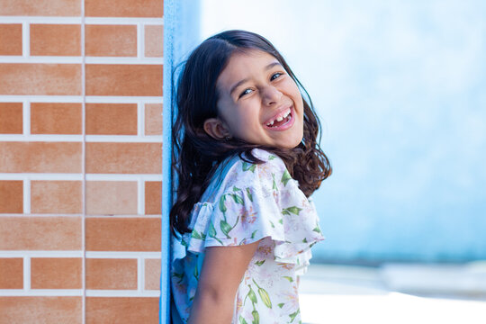 Uma criança, menina, brasileira dos cabelos pretos e lisos, alegre, sorrindo, encostada no muro.