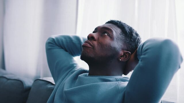 Slow-motion footage of a serious man thinking while sitting on a blue sofa. Hands behind the head and looking at the ceiling of the room. Focused. Cozy room with white curtains in the background. 