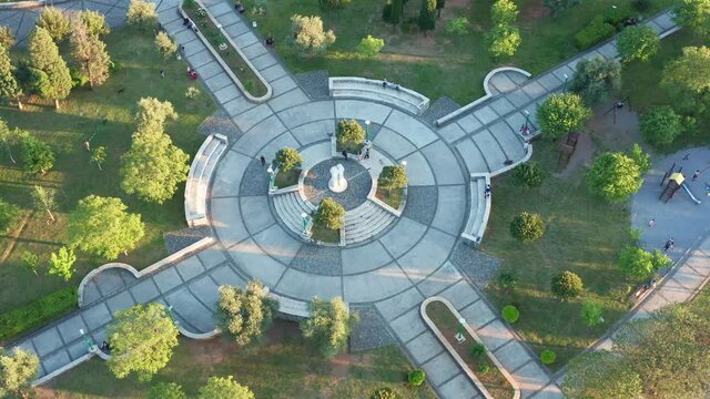 Round paved playground in a garden surrounded by trees and grass, as the central square of green city park and recreational area in urban environment. Aerial drone view of walkways and podium.
