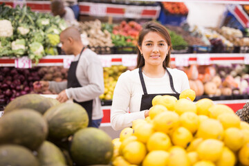 Latin saleswoman in black apron and her assistant with box of fruits in background