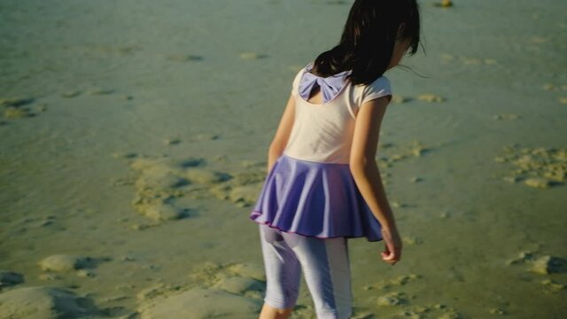 A long black-haired Asian girl in a purple swimming suit and barefoot is playing at beach on summer vacation. By writing and drawing on the wet sand at the time of the sunset on beautiful island.