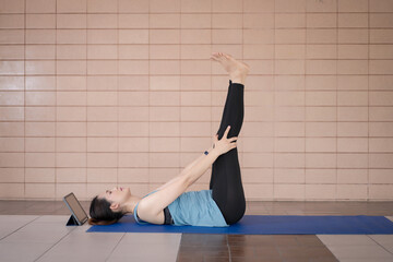 A middle-aged Asian woman in relaxed sports outfits doing an exercise workout training program follows an online workout lesson on a tablet at home during the COVID-19 pandemic and city lockdown