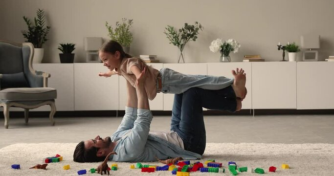 Caring strong young father lying on carpet, lifting in air little cute daughter, having fun together in living room. Laughing bonding 30s man and small kid girl playing airplane, imagining travelling.