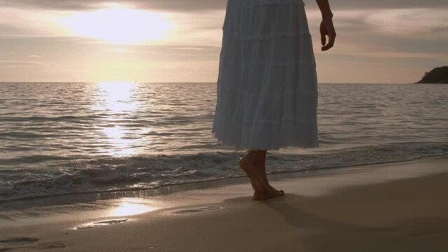 4K Slow motion woman feet walking barefoot on the beach at golden sunset time. leaving footprints in sand. Female tourist on summer vacation holiday relax at tropical beach Phuket, Thailand. 