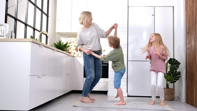 Excited mom or babysiter and two kids boy and girl jumping, dancing laughing in modern scandinavian house kitchen. Happy family mother daughter son having fun enjoy playing singing together at home.
