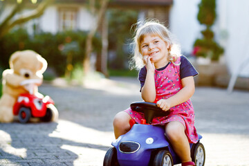 Little adorable toddler girl driving toy car and having fun with playing with plush toy bear, outdoors. Gorgeous happy healthy child enjoying warm summer day. Smiling stunning kid in gaden
