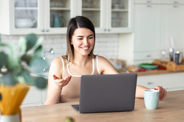Young woman is using laptop and working sitting at the table at home in the kitchen. Beautiful female communicating on social networks using computer