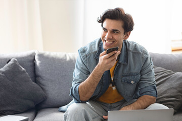 Young successful freelance entrepreneur working remotely at home using laptop and smartphone. Man talking on the phone while sitting on the couch at home