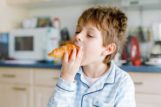 Happy little blond kid boy eating fresh croissant for breakfast or lunch. Healthy eating for children. Child in colorful pajama sitting in domestic kitchen after sleeping in the morning.