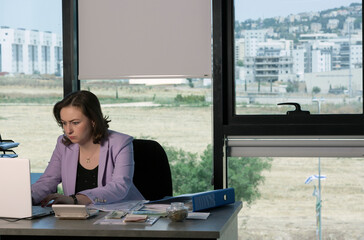 Woman thinking about something in Office, working using laptop reading email from client. Dollars, shekels Money on the table. Saving money, long hours of work, taxes, business finance and accounting