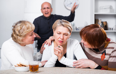Mature woman is sitting upset and her girlfriends are expressioning understanding to she at home.