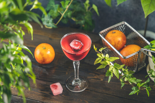 Elegant stemware glass of blood orange cocktail with frozen raspberries in ice cube on dark wooden table surface